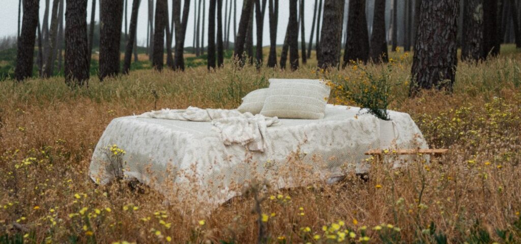Cama decorada en tonos neutros en medio de un campo florido con árboles al fondo, reflejo de elegancia y confort.