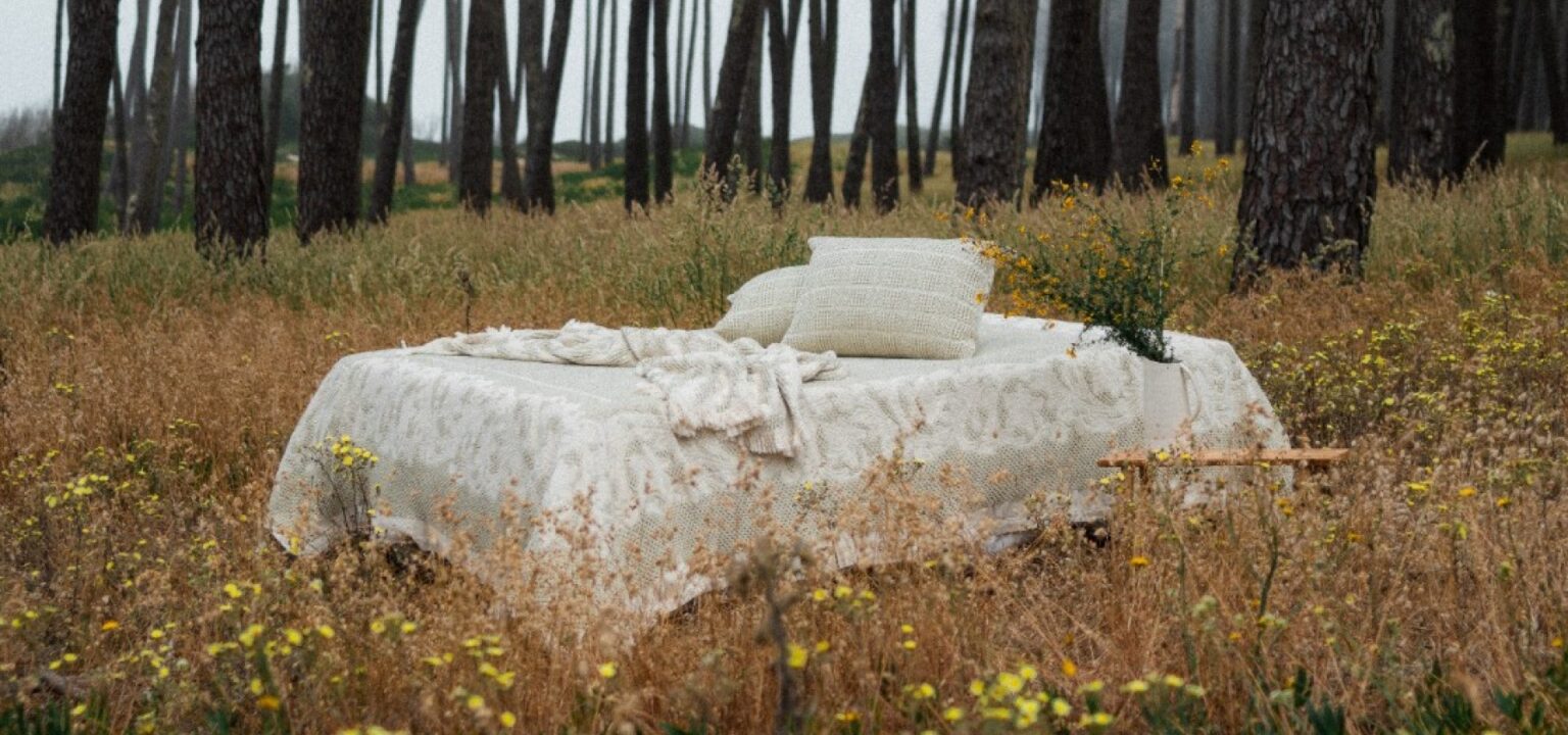 Cama decorada com tons neutros em meio a um campo florido com árvores ao fundo, refletindo elegância e conforto.