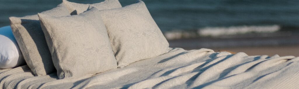 Cama con cojines y colcha texturizada en colores claros, situada frente al mar, que representa el confort y la sofisticación.