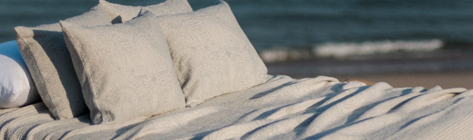 Cama com almofadas e colcha texturizada em tons claros, posicionada em frente ao mar, representando conforto e sofisticação.