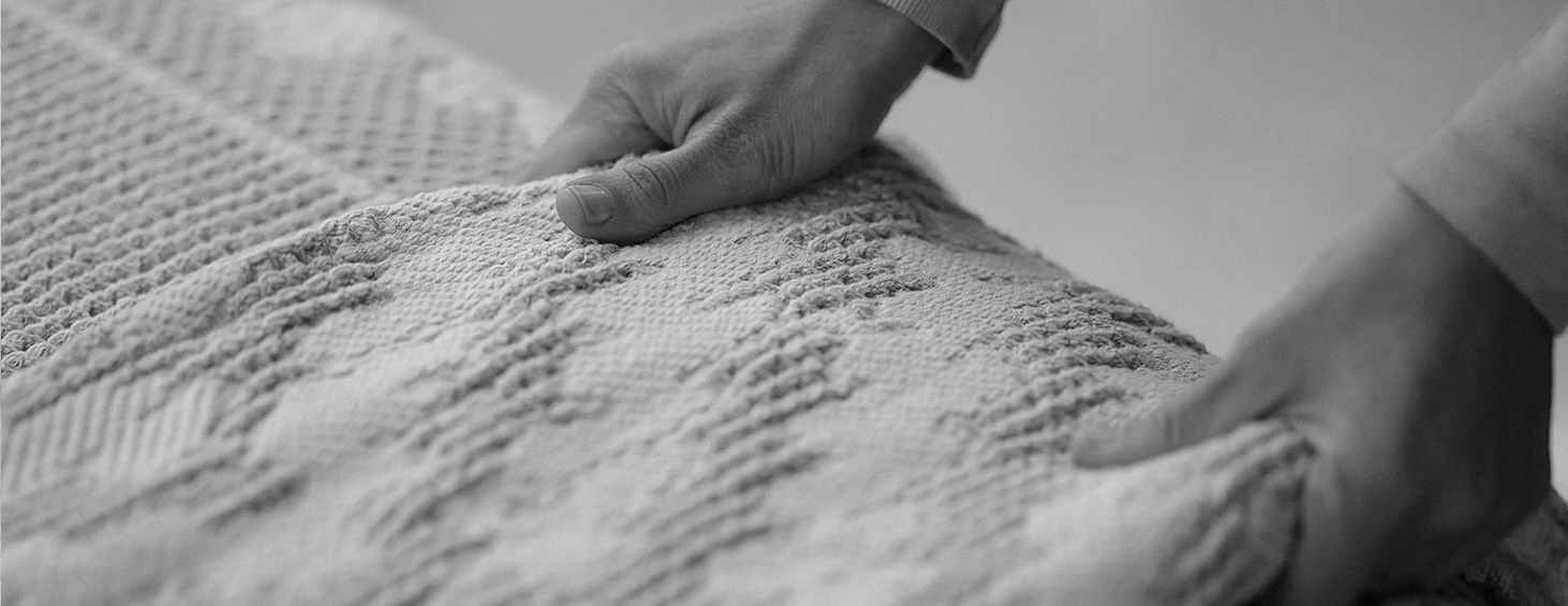 Mãos ajustando uma colcha texturizada em close-up, destacando a suavidade e os detalhes de qualidade dos produtos Cama em Ponto.